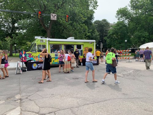A colorful food truck at a street event, with people gathered around and trees in the background.