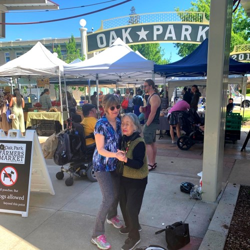 Two women smile and embrace at a bustling farmers market, surrounded by tents and shoppers in Oak Park.
