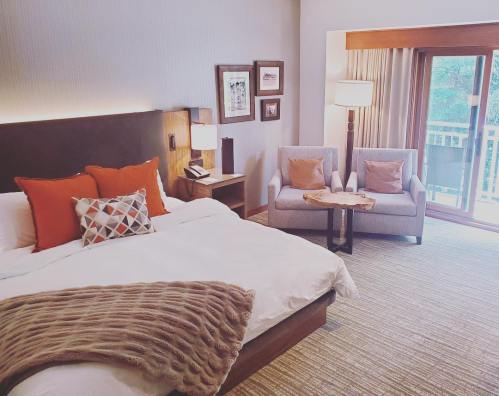 Cozy hotel room featuring a king bed, two armchairs, a wooden table, and a balcony with natural light.