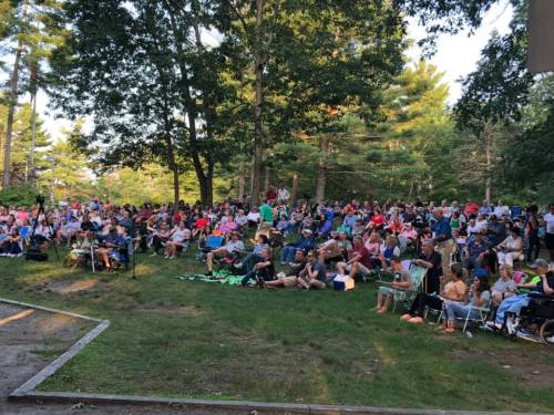 A large crowd of people seated on the grass in a park, enjoying an outdoor event surrounded by trees.