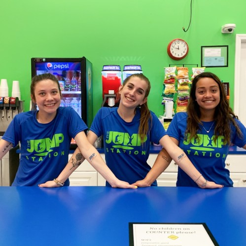 Three smiling young women in matching blue shirts stand behind a counter, with snacks and drinks in the background.
