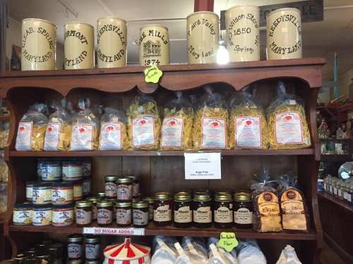 A wooden shelf displays jars, pasta, honey, and flour, showcasing local products in a rustic store setting.