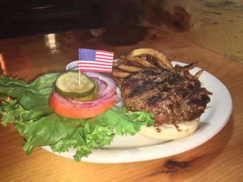 A grilled burger on a bun with lettuce, tomato, pickles, onion, and fries, topped with a small American flag.