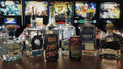 A selection of various liquor bottles and a can of beer on a bar, with pinball machines in the background.