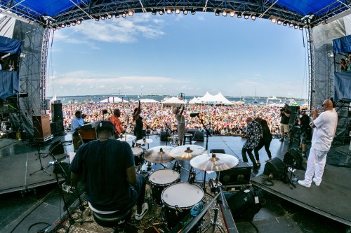 A lively outdoor concert scene with a large crowd, musicians on stage, and a clear blue sky in the background.