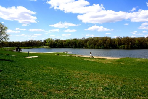 A serene landscape featuring a grassy area by a calm lake, with trees and fluffy clouds in a blue sky.