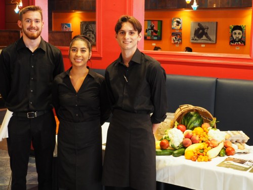 Three restaurant staff members in black uniforms stand smiling beside a display of fresh vegetables and eggs.