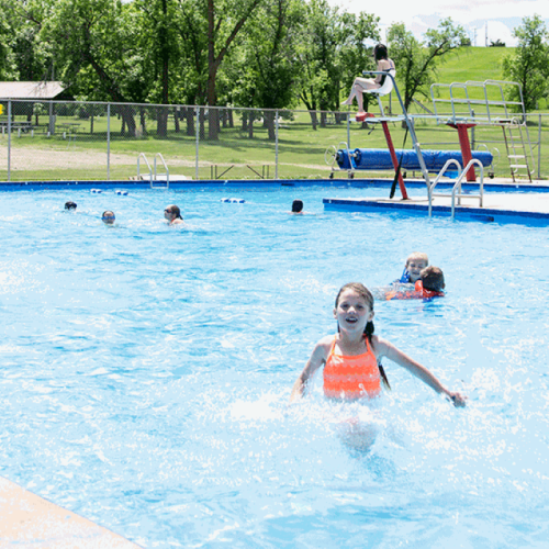 A girl in an orange swimsuit splashes in a pool while other children swim and play nearby on a sunny day.