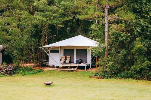 A spacious tent cabin in a wooded area, with a deck and two chairs, surrounded by greenery and a fire pit nearby.