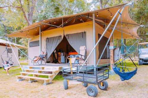A glamping tent with a porch, chairs, and a cart outside, surrounded by trees in a natural setting.