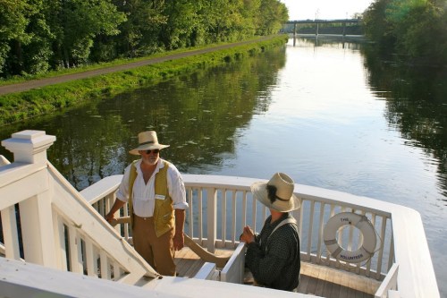 Two people in historical attire stand on a boat by a calm river, surrounded by lush greenery and a bridge in the background.