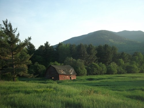 A weathered red barn sits in a lush green field, surrounded by trees and mountains under a clear sky.