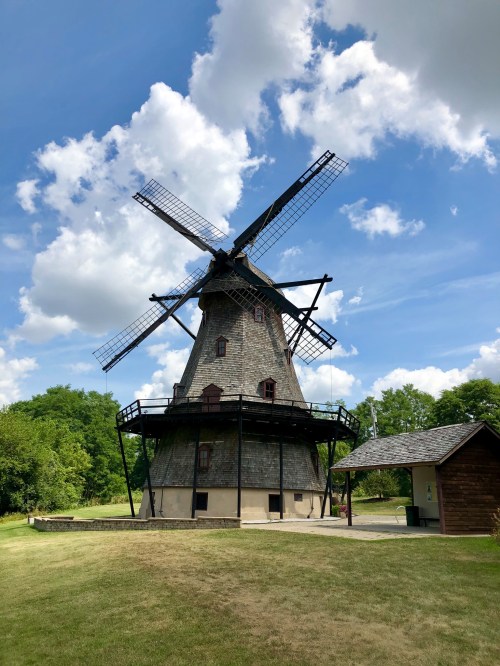 A historic windmill with large blades stands against a blue sky with fluffy clouds, surrounded by green trees.