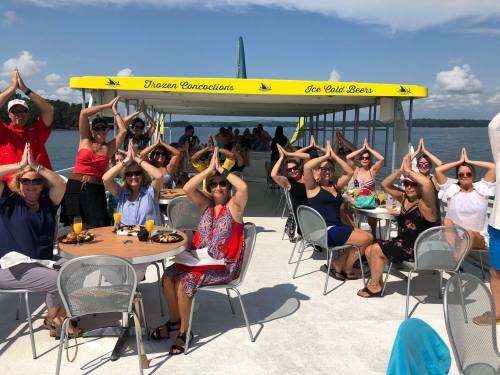 A group of people on a boat, smiling and posing with hands raised in a playful gesture, enjoying a sunny day.