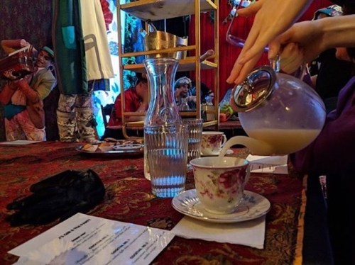 A hand pours a drink from a glass pitcher into a floral teacup, with a table set for tea in a vibrant, colorful setting.