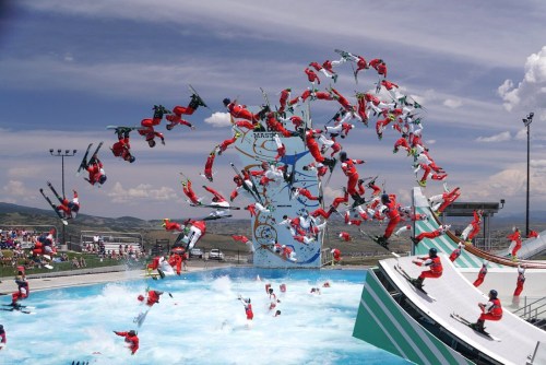 A dynamic scene of skiers performing tricks above a water ramp, with spectators watching in the background.