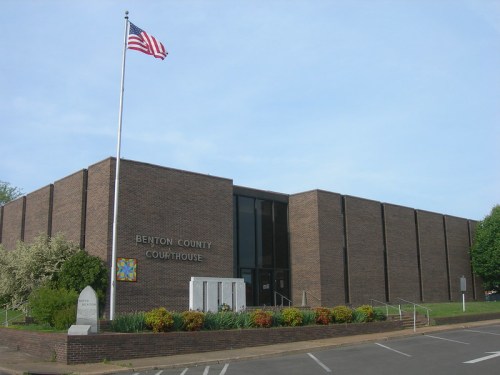 Benton County Courthouse with American flag, surrounded by greenery and flowers on a clear day.