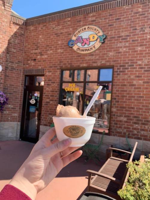 A hand holds a cup of ice cream in front of a brick building with a sign for a candy company.