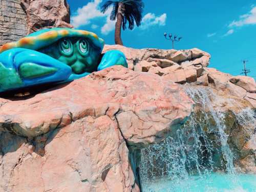 A colorful crab sculpture rests on rocks near a waterfall, with a palm tree and blue sky in the background.