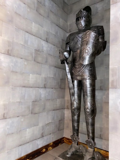 A tall, metallic knight statue stands in a corner, wearing armor and holding a sword, against a gray stone wall.