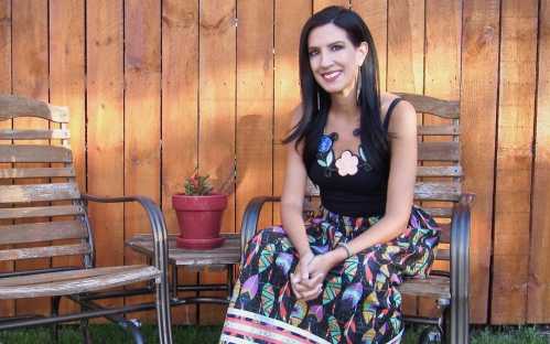 A woman in a colorful skirt sits on a chair beside a potted plant, smiling against a wooden fence backdrop.