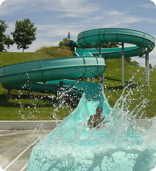 A person slides down a turquoise water slide, splashing into the pool below, with green hills in the background.