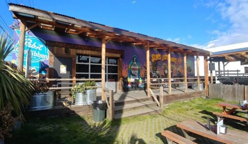 A colorful building with a mural, wooden deck, and outdoor seating, surrounded by greenery and blue skies.