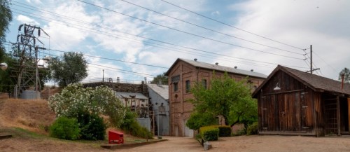 A panoramic view of an industrial site with power lines, brick buildings, and greenery under a cloudy sky.