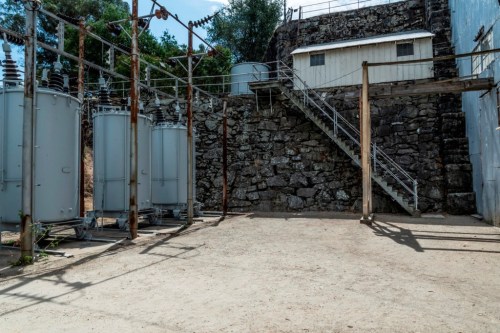 Industrial area with transformers, a stone wall, and a staircase leading to a building in the background.