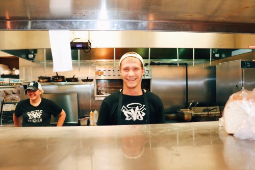 Two young men in a kitchen, one smiling at the camera while the other stands behind him, both wearing black shirts.
