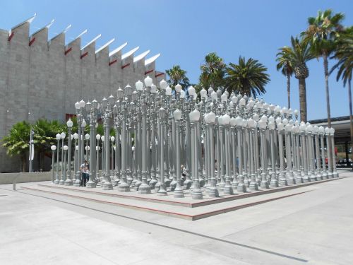 A large installation of vintage street lamps in a sunny outdoor space, surrounded by palm trees and modern architecture.