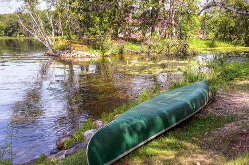 A green canoe rests on the grassy shore beside a calm lake surrounded by trees and lily pads.