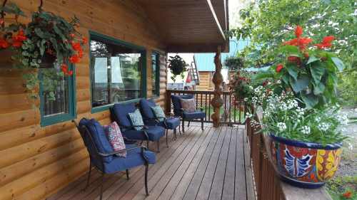 A cozy wooden porch with blue chairs, colorful flower pots, and hanging plants, surrounded by greenery.