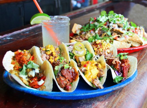 A vibrant platter of tacos with various fillings, accompanied by a drink and a large plate of nachos in the background.