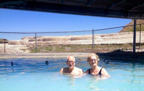 Two people smiling in a pool under a clear blue sky, with a fenced area and hills in the background.