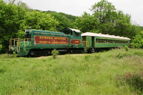 A green vintage train with "Eureka Springs North Arkansas" on the side, parked amidst lush greenery.