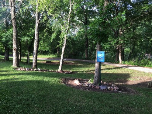 A serene park scene with trees, a grassy area, and a sign near a winding path. Sunlight filters through the leaves.