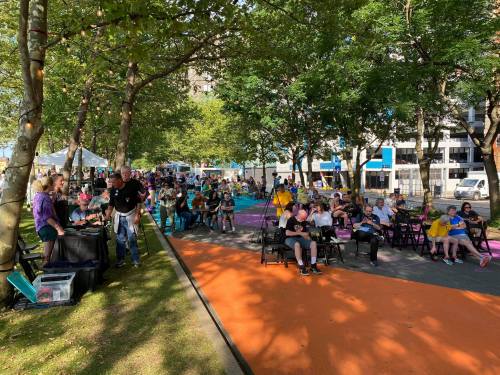 A lively outdoor event with people seated in chairs under trees, enjoying the atmosphere in a colorful park setting.