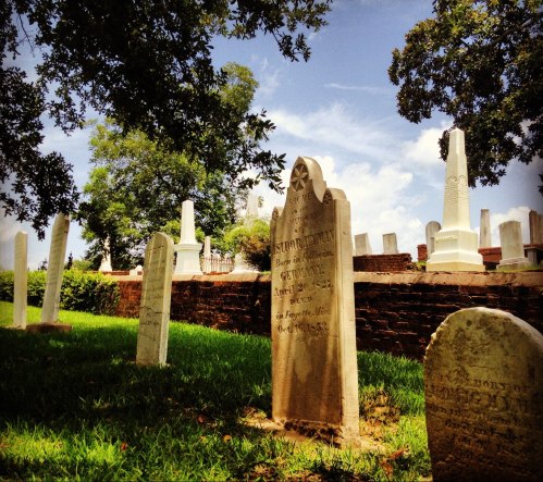 A peaceful cemetery scene with gravestones surrounded by green grass and trees under a blue sky.