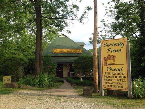 A rustic building with a green roof and a sign reading "Serenity Farm Bread," surrounded by trees and greenery.