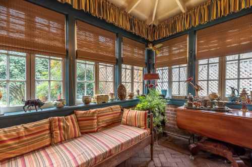 Cozy sunroom with striped sofa, plants, and decorative items, featuring large windows and warm, inviting decor.