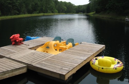A wooden dock on a calm lake with a yellow paddle boat and a colorful inflatable tube nearby. Green trees line the shore.