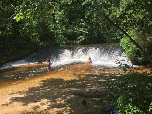 Two people play in a shallow river near a small waterfall, surrounded by lush green trees and sunlight.