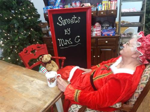 A woman in a red Santa outfit sits at a table with a teddy bear, next to a sign reading "Sweets by Mrs. C."