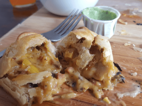 A close-up of a flaky pastry filled with cheese, corn, and meat, served with a small container of green sauce.
