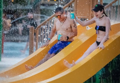 A man and a woman slide down a yellow water slide, enjoying drinks as water splashes around them.