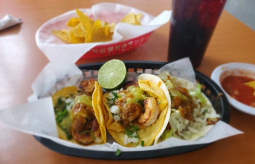Three tacos filled with shrimp, lettuce, and salsa, served with a lime wedge, tortilla chips, and a drink in the background.