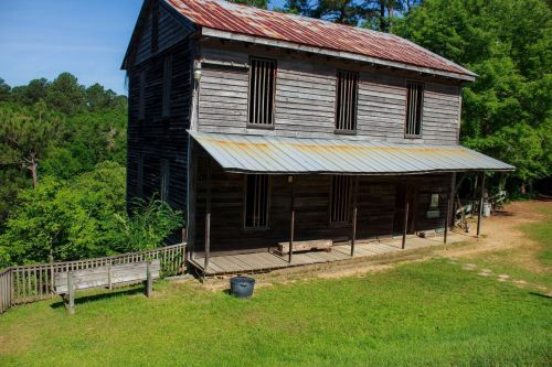 A weathered wooden building with a metal roof, surrounded by greenery and featuring a porch and benches.