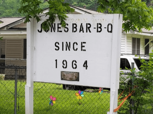 Sign for Jones Bar-B-Q, established in 1964, in front of a house with colorful decorations in the yard.