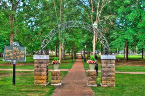 Archway entrance to a park with a pathway lined by trees, featuring a historical marker on the left.